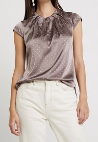 Satin blouse met korte mouwen in mocha met witte stippen. Heeft een geraffineerde halslijn en een losvallende pasvorm, gecombineerd met lichtgekleurde jeans.