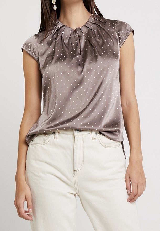 Satin blouse met korte mouwen in mocha met witte stippen. Heeft een geraffineerde halslijn en een losvallende pasvorm, gecombineerd met lichtgekleurde jeans.