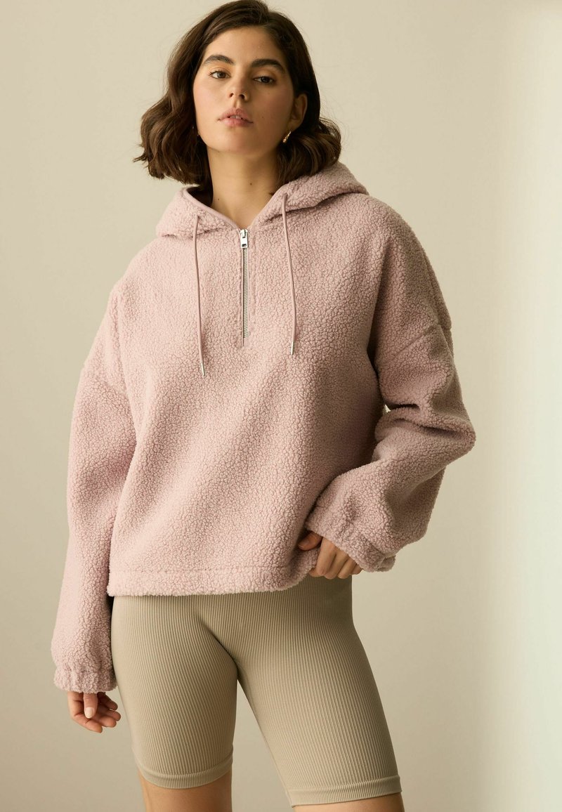 Roze fleece hoodie met een halve rits, ontspannen pasvorm en oversized mouwen, gecombineerd met beige geribbelde shorts. Zachte textuur en casual ontwerp.