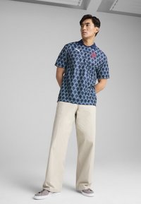Polo shirt bleu marine avec un motif géométrique bleu, manches courtes, et un logo rouge, associé à un pantalon beige et des baskets grises.