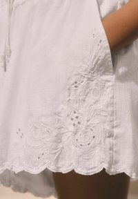 Vitt linneplagg med scalloped fåll som har blommönster broderat och utklippta mönster. Mjuk textur och avslappnad design.
