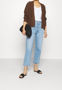 Brauner gestrickter Cardigan über einem weißen T-Shirt, kombiniert mit hellblauen geraden Jeans und schwarzen Rutsch-Sandalen. Schwarze strukturierte Handtasche.
