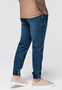 Pantalones jogger de denim azul con puños elásticos, bolsillos delanteros y traseros, usados con zapatillas beige. Textura suave, ajuste entallado, estilo casual.