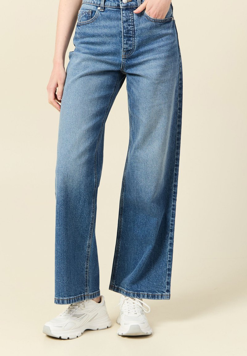 Jasnoniebieskie dżinsy o szerokich nogawkach z wysokim stanem, subtelnym efektem przetarcia i kontrastowymi szwami, zestawione z białymi trampkami.