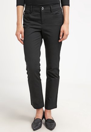 Kobieta ubrana w dopasowane czarne spodnie z przednimi kieszeniami i suwakiem, w połączeniu z czarnymi teksturowanymi butami na płaskim obcasie o szpiczastym czubku.