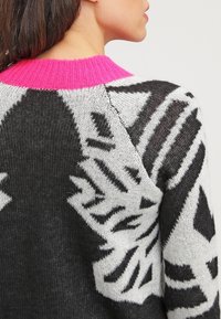 Nainen, joka käyttää mustavalkoista kuviollista neulepaitaa, jossa on kirkkaan vaaleanpunainen ribbikaulus. Hänet on kuvattu takaapäin ja hieman kääntyneenä kyljelleen.