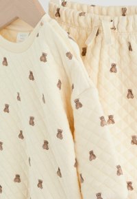 Quiltet baby outfit i blød cremefarvet stof med et bamsemotiv. Sættet inkluderer en langærmet top og bukser med elastik i taljen.