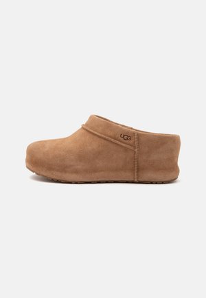 Bruine suède UGG slip-on klomp met lage hak en afgeronde neus, weergegeven in zijaanzicht op een witte achtergrond.