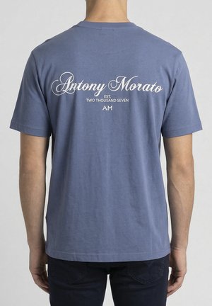 Uomo che indossa una maglietta blu a maniche corte con testo in corsivo bianco sul retro: "Antony Morato EST. TWO THOUSAND SEVEN AM".