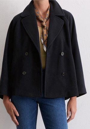Person trägt einen doppelt geknöpften schwarzen Mantel über einem gelben Top, blauen Jeans und einem braun gemusterten Halstuch, vorne gebunden.