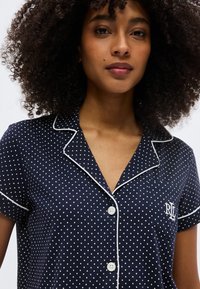 Νεαρή γυναίκα με σγουρά μαλλιά που φοράει ένα μπλε ναυτικό πουκάμισο με λευκές ρίγες και πουά, με κεντημένο το γράμμα "RL" στην τσέπη στο στήθος.