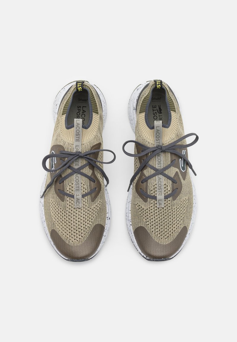Zapatillas de punto beige con detalles marrones, punta redonda, cordones grises y suela blanca moteada. Presenta la marca Lacoste en la lengüeta.
