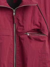 Geacă roșie ușoară, cu închidere frontală cu fermoar, buzunar lateral cu fermoar și țesătură netedă, texturată. Accentele includ detalii negre.