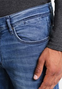 Blå denimjeans med en klassisk design som har fem fickor, kontrasterande sömmar och en lätt tvättad effekt, visade mot en grå topp.