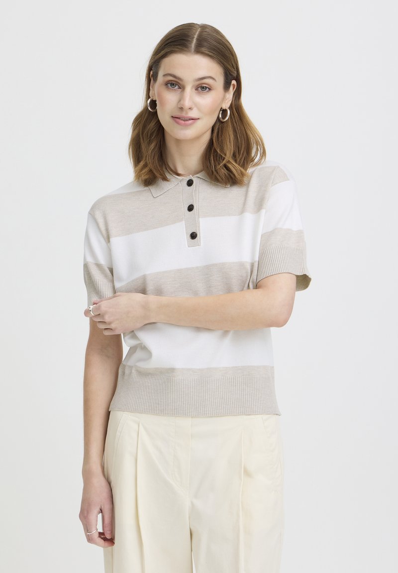 Femme aux cheveux bruns mi-longs portant un polo à manches courtes beige et blanc rayé et un pantalon plissé crème, debout les bras croisés.