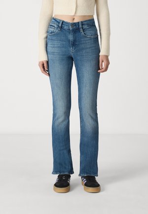 Jean bleu évasé en denim, avec une taille mi-haute et une décoloration subtile. Associé avec des baskets noires.