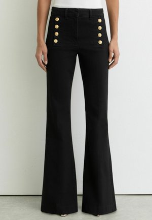 Pantalon évasé noir taille haute avec deux rangées de boutons dorés sur les poches avant, porté avec un haut blanc.
