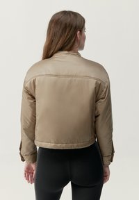 Tan beskuren pufferjacka i glänsande material med ståkrage, elastiska ärmslut och detalj i ryggen. Matchas med svarta texturerade leggings.
