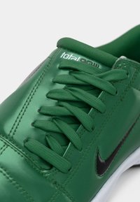 Zöld sportcipő sima szintetikus felsőrészsel, textil fűzőkkel, fekete Nike logóval és fehér gumitalppal. Total90 márkajelzés a nyelvnél.