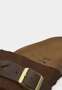 Καφέ δερμάτινο σανδάλι με ρυθμιζόμενο χρυσό πόρπη και πέλμα από σουέτ, κοντινή λήψη που δείχνει την υφή του λουριού και το ανάγλυφο λογότυπο εσωτερικά.