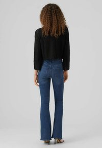 Pull cropped en tricot noir avec motif texturé, associé à un jean flare bleu taille haute, vu de dos. Talons aiguilles visibles.