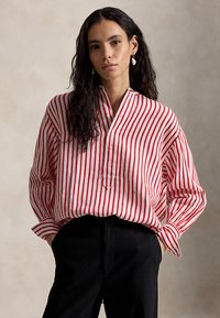 Žena s dlhými tmavými vlasmi, oblečená v červeno-bielej blúzke s vertikálnymi pruhmi a čiernych nohaviciach, stojaca pred jednofarebným svetlým pozadím.