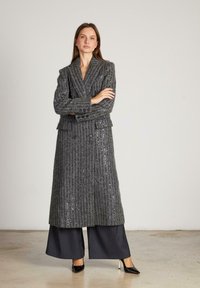 Abrigo gris, largo, de doble botón con rayas verticales y textura de lentejuelas. Lleva puesto sobre pantalones oscuros de pierna ancha y zapatos de tacón alto.