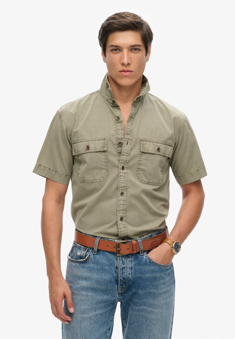 Camisa de manga corta color oliva con botones y dos bolsillos en el pecho, combinada con jeans azules desgastados y un cinturón de cuero marrón. Reloj en la muñeca.