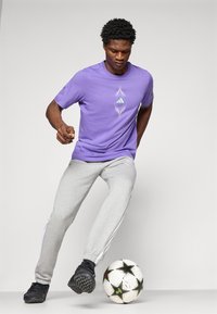Lilla bomulds t-shirt med grafisk design, parret med grå sweatpants med sideløsninger og sorte fodboldsko. En fodbold ligger på jorden.