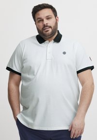Bijela majica s kratkim rukavima s crnim ovratnikom i detaljima na rukavima. Sadrži plastron s tri gumba i mali logo globusa na prsima.