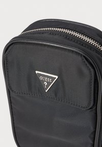 Svart nylonveske med et trekantet sølvlogo, med glidelås og en strukturert overflate. Enkel, geometrisk design.