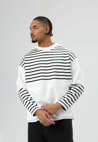 Witte sweater met lange mouwen en zwarte horizontale strepen over de borst en mouwen. Verkrijgbaar met een ronde hals en een comfortabele pasvorm.