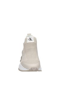 Světlé béžové sportovní tenisky s pleteným svrškem, výrazná logo visačka na jazyku a strukturovaná podrážka. Obsahují poutko na zadní straně.