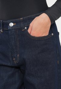 Ciemnoniebieskie dżinsy o dopasowanym kroju, z widocznym przeszyciem, mosiężnym guzikiem i przednią kieszenią. Materiał ma gładką fakturę i subtelny połysk.