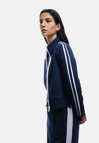Chaqueta de cremallera azul marino con rayas blancas en los costados, cuello alto y dobladillos acanalados. Confeccionada en un tejido suave y liso con un ajuste estructurado.