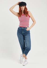 Rood-witte gestreepte tanktop, high-waisted blauwe spijkerbroek en witte canvas sneakers. Model poseert met één hand op de heup, terwijl ze een zwarte pet draagt.