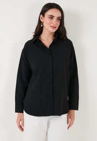 Black button-up shirt with vertical ribbed texture, long sleeves, and a classic collar. Paired with white trousers, featuring black buttons.