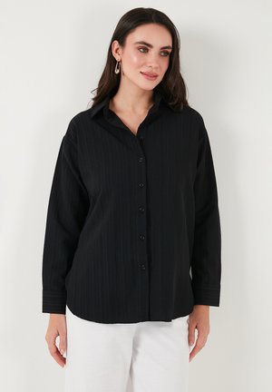 Black button-up shirt with vertical ribbed texture, long sleeves, and a classic collar. Paired with white trousers, featuring black buttons.