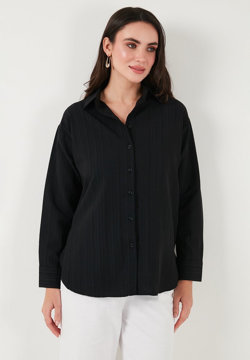 Black button-up shirt with vertical ribbed texture, long sleeves, and a classic collar. Paired with white trousers, featuring black buttons.
