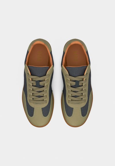 Ζευγάρι αθλητικών παπουτσιών με κορδόνια σε ελιά πράσινο και μαύρο χρώμα, με πορτοκαλί εσωτερική επένδυση και καφέ σόλες, φωτογραφημένα από πάνω σε λευκό φόντο.