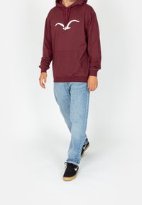 Felpa cappuccio bordeaux con logo bianco, tasca frontale e vestibilità comoda, abbinata a jeans azzurri chiari e sneakers nere.