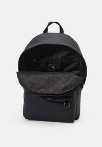 Mochila azul marino con un exterior liso, cierre de cremallera y un bolsillo interior. Forrada con una tela estampada que presenta un sutil diseño de logo.