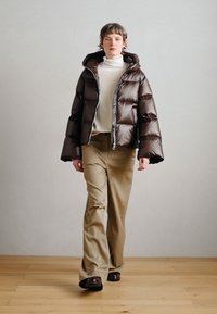 Pruun läikiva viimistlusega tepitud jope, millel on ees tõmblukk ja suur kapuuts, kantud valge kõrge kaelusega kampsuni ja beežide laia jala pükstega.