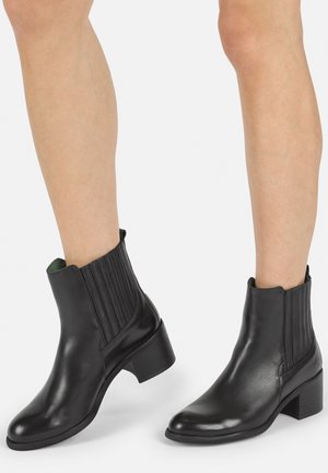 Botas de tobillo de cuero negro con un acabado liso, paneles laterales elásticos y un tacón bajo en bloque. Presentan una puntera redondeada y detalles de costura sutiles.