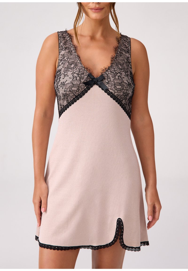 Kobieta w beżowej, bezrękawnikowej koszuli nocnej z czarną koronkową lamówką na biuście, dekoltem w kształcie litery V, czarną kokardką na środku i falowanym dołem z niewielkim przednim rozcięciem.