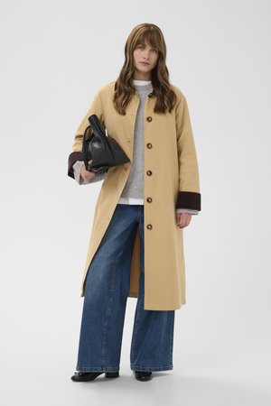 Mujer con un abrigo largo beige con puños oscuros, suéter gris, vaqueros anchos azules, zapatos negros, llevando un bolso negro con textura.