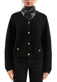 Frau trägt eine schwarze strukturierte Strickjacke mit goldenen Knöpfen, schwarze Hose und einen schwarzen Lederschal um den Hals vor einem weißen Hintergrund.
