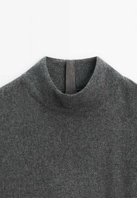 Pull à col roulé en laine gris foncé avec une étiquette boucle en tissu noir au centre du dos du col sur fond blanc.