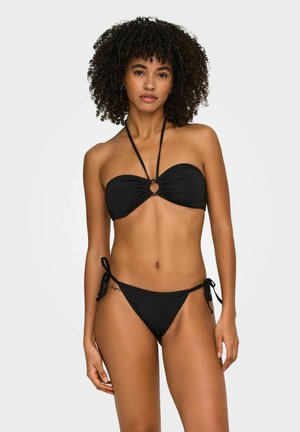 Woman with curly hair wearing a black strapless bikini top with halter straps and matching black bikini bottoms with side ties.
