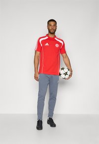 Red short-sleeve sports shirt with white accents and logo, paired with light gray tapered sweatpants. Holding a soccer ball. Black sneakers.
